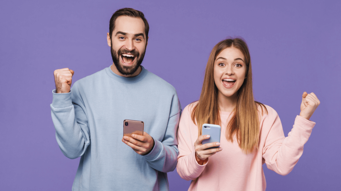 Uśmiechnięty mężczyzna i kobieta w pastelowych bluzach, trzymający smartfony i unoszący zaciśnięte pięści w geście radości na tle fioletowej ściany.